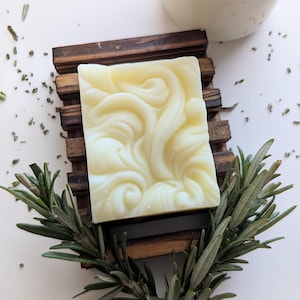 May include: A square, off-white bar of soap with a raised, swirling, embossed pattern on its surface. The soap rests on a dark brown, slatted wooden soap dish, accompanied by a sprig of fresh green rosemary and a glass jar with a cork lid.