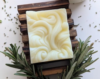 May include: A square, off-white bar of soap with a raised, swirling, embossed pattern on its surface. The soap rests on a dark brown, slatted wooden soap dish, accompanied by a sprig of fresh green rosemary and a glass jar with a cork lid.