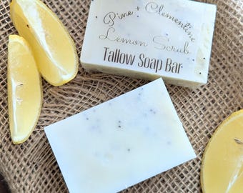 Könnte beinhalten: Zwei weiße Seifenstücke mit kleinen dunklen Punkten, beschriftet mit "Pine + Clementine Lemon Scrub Tallow Soap Bar", neben Zitronenspalten. Ein Glas mit Deckel befindet sich im Hintergrund, alles auf einer Jutefläche.