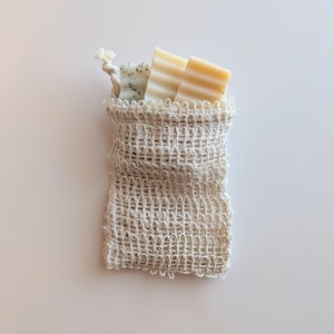 Puede incluir: Una bolsa de jabón tejida natural llena de tres pastillas de jabón. Una pastilla está moteada con semillas negras, y las otras dos son de color amarillo claro con líneas horizontales. La bolsa está atada en la parte superior con un cordón anudado.