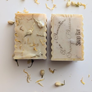 May include: Two rectangular soap bars with a light beige hue, adorned with dried flower petals and buds. One bar is labeled "Pine + Clementine Orange Blossom Soap Bar". The soaps are arranged on a white surface, with scattered petals.