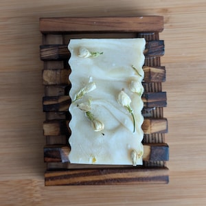 Puede incluir: Una pastilla rectangular de jabón artesanal con borde ondulado, sobre una jabonera de madera. El jabón es de color claro con remolinos blancos y está decorado con pequeños capullos de flores blancas. La jabonera es de madera marrón oscuro.