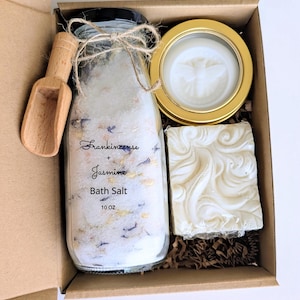 Puede incluir: Un juego de regalo en una caja de cartón que contiene una botella de vidrio de sal de baño, una cuchara de madera, una vela dorada en una lata y una pastilla de jabón. La botella de sal de baño está etiquetada como "Frankincense + Jasmine Bath Salt 283g."