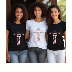 May include: Three women wearing t-shirts with the word "MARZIPAN" in pink script. The shirts are black and white, and feature a pink ballet shoe design. The women are smiling and wearing jeans.
