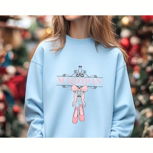 May include: A light blue crewneck sweatshirt featuring a graphic print of a black decorative scrollwork frame with the word MARZIPAN in pink letters, accompanied by a pair of pink ballet slippers tied together with a matching pink ribbon bow.