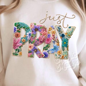 May include: Cream-coloured sweatshirt featuring the words "just pray" in a floral design. The word "pray" is spelled out in colourful flowers. The word "just" is written in cursive above the word "pray".