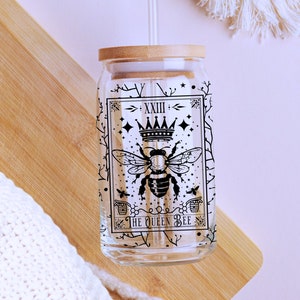 May include: A clear glass reusable water bottle with a bamboo lid and a black and white illustration of a bee with a crown and the text "The Queen Bee" on the side. The bottle is sitting on a wooden surface.
