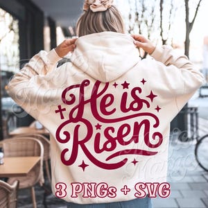 May include: Cream-colored hoodie with the phrase "He is Risen" in burgundy script on the back. The design includes crosses and stars. The bottom of the hoodie reads "3 PNGs + SVG".