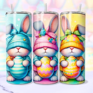 Op de afbeelding: Drie roestvrijstalen bekers met kleurrijke paasbunny-ontwerpen. Elke bunny draagt ​​een hoed en houdt een paasei vast. De bunnies zijn blauw, roze en geel.