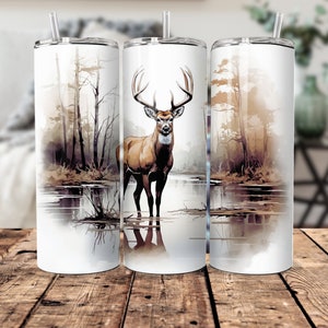 May include: A white stainless steel tumbler with a watercolor design of a deer standing in a lake with trees in the background.