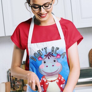 May include: A white apron with a blue background and a cartoon hippopotamus wearing a red and white shirt. The text "Don't be a hippopotamus" is printed on the apron. A person is wearing the apron and holding a knife while cutting a tomato.