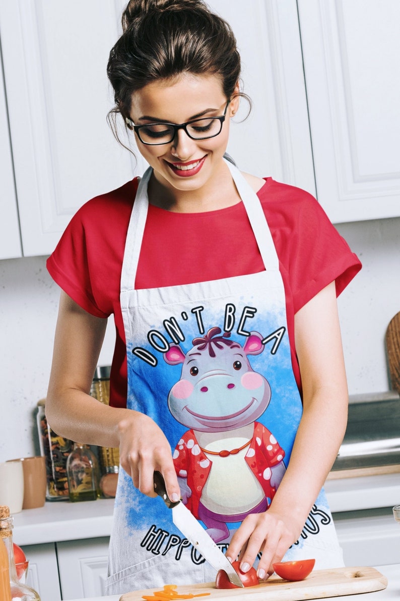 May include: A white apron with a blue background and a cartoon hippopotamus wearing a red and white shirt. The text "Don't be a hippopotamus" is printed on the apron. A person is wearing the apron and holding a knife while cutting a tomato.