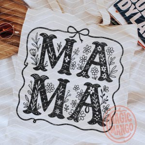 Puede incluir: Una bolsa de tela blanca con un dise&ntilde;o en blanco y negro que dice "MAMA" con detalles florales. El dise&ntilde;o est&aacute; enmarcado en un borde negro con un peque&ntilde;o lazo en la parte superior. La bolsa tiene un logotipo de "Magic Mango" en la esquina inferior derecha.