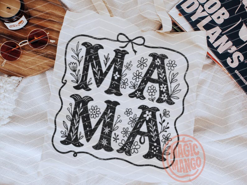 Puede incluir: Una bolsa de tela blanca con un dise&ntilde;o en blanco y negro que dice "MAMA" con detalles florales. El dise&ntilde;o est&aacute; enmarcado en un borde negro con un peque&ntilde;o lazo en la parte superior. La bolsa tiene un logotipo de "Magic Mango" en la esquina inferior derecha.