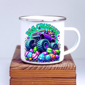 May include: White enamel mug featuring a purple monster truck carrying Easter eggs. The truck is driving over a pile of colourful Easter eggs. The text "Egg Crusher" is printed on the mug in green and white.