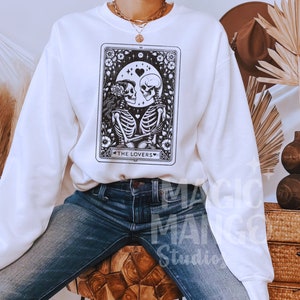 May include: White sweatshirt featuring a black and white illustration of a tarot card depicting two skeletons holding hands. The card is labelled "The Lovers".