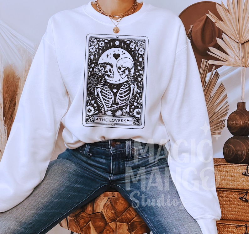 May include: White sweatshirt featuring a black and white illustration of a tarot card depicting two skeletons holding hands. The card is labelled "The Lovers".