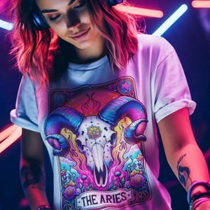 May include: White t-shirt with a colourful graphic of a ram skull and the text "THE ARIES". The design incorporates blues, purples, pinks, and yellows. The shirt has short sleeves and a crew neck.