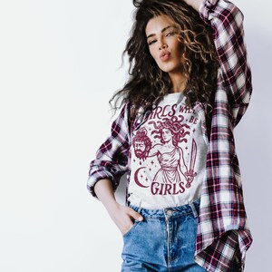 May include: A light gray t-shirt with a burgundy graphic of Medusa holding a sword and the severed head of a man. The text "GIRLS WILL BE GIRLS" is above and below the image. The person is wearing a plaid shirt.
