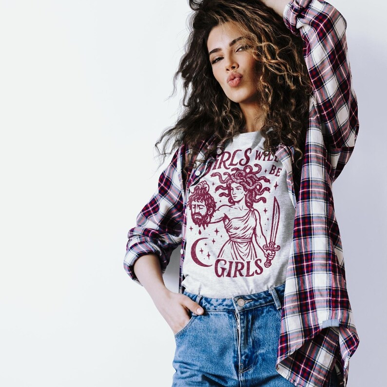 May include: A light gray t-shirt with a burgundy graphic of Medusa holding a sword and the severed head of a man. The text "GIRLS WILL BE GIRLS" is above and below the image. The person is wearing a plaid shirt.