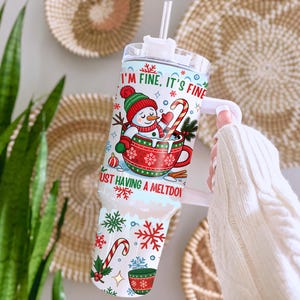 May include: A white tumbler with a handle, featuring a winter-themed design. The design includes a snowman in a red cup, candy canes, and snowflakes. Text on the tumbler reads "I'm fine, it's fine" and "Just having a meltdown".