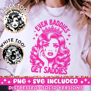 May include: A white t-shirt with a pink graphic design featuring a woman with a crown and the text "Even Baddies Get Saddies". The graphic is surrounded by stars and the text "Black Too!" and "White Too!" are in the corners of the image.