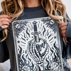 Pu&ograve; includere: Felpa grigio scuro con grafica in bianco e nero. La grafica raffigura una mano scheletrica che tiene un cubo trafitto da un pugnale, circondato da fiamme, stelle ed elementi floreali. Il design ha un'estetica vintage e gotica.