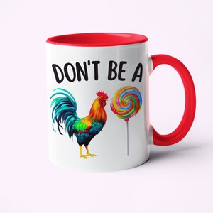 May include: White ceramic coffee mug with a red rim and handle. The mug features a colorful rooster illustration and a lollipop with the text "DON'T BE A" printed in black.