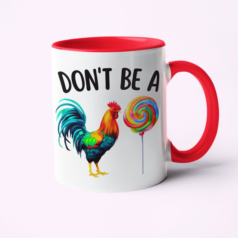May include: White ceramic coffee mug with a red rim and handle. The mug features a colorful rooster illustration and a lollipop with the text "DON'T BE A" printed in black.