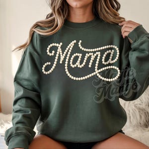 May include: A dark green sweatshirt with the word "Mama" in a cursive font, outlined with small, off-white pearls. The sweatshirt has long sleeves and a crew neck. The brand name "Marie Mango" is printed at the bottom right.