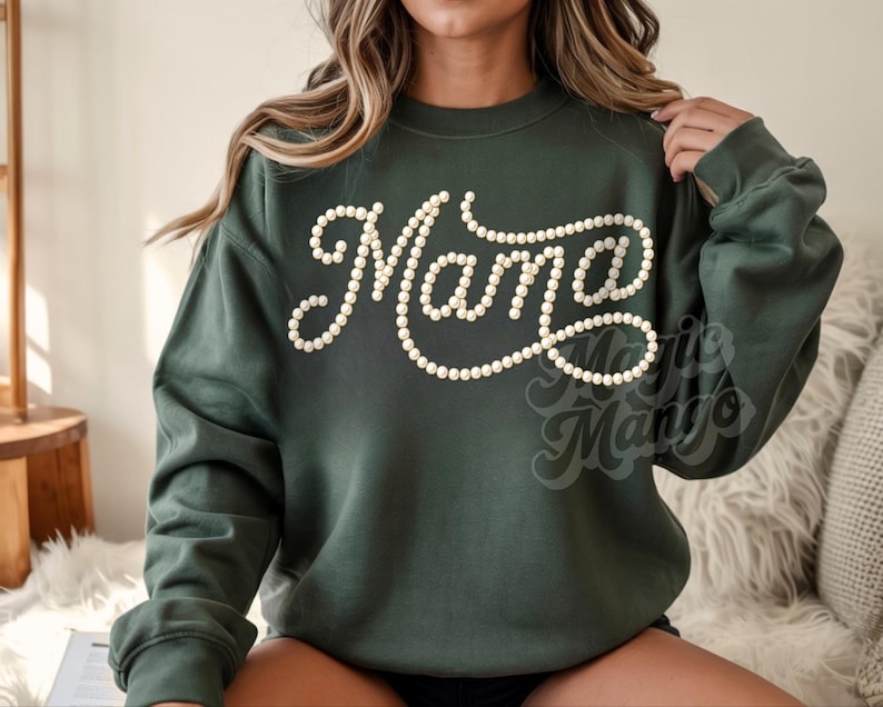 May include: A dark green sweatshirt with the word "Mama" in a cursive font, outlined with small, off-white pearls. The sweatshirt has long sleeves and a crew neck. The brand name "Marie Mango" is printed at the bottom right.