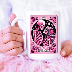 K&ouml;nnte beinhalten: Wei&szlig;er Keramikbecher mit einem rosa Tarotkarten-Design. Die Karte zeigt Skelettfiguren und tr&auml;gt die Aufschrift "AMOR" und "THE LOVERS". Das Design umfasst florale Elemente und ein Herz.
