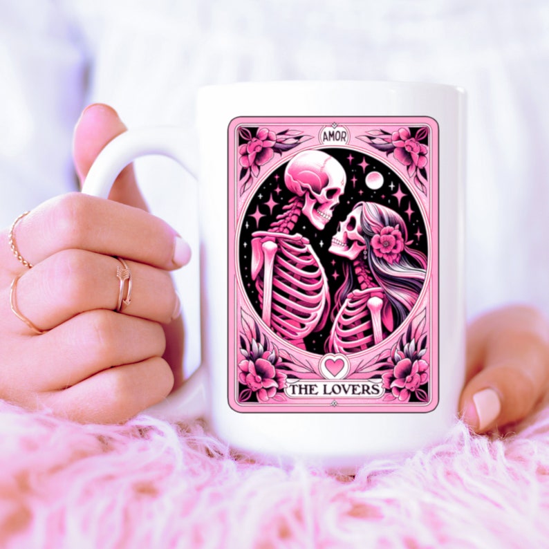 K&ouml;nnte beinhalten: Wei&szlig;er Keramikbecher mit einem rosa Tarotkarten-Design. Die Karte zeigt Skelettfiguren und tr&auml;gt die Aufschrift "AMOR" und "THE LOVERS". Das Design umfasst florale Elemente und ein Herz.