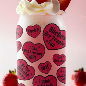 Puede incluir: Un vaso con una bebida rosa, cubierto con crema batida y rodajas de fresa. El vaso est&aacute; decorado con pegatinas rosas en forma de coraz&oacute;n con texto negro, incluyendo frases como "Fuck It" y "Bitches Are People Too."