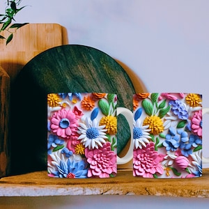 May include: Two white ceramic mugs with a colorful floral design. The flowers are pink, blue, yellow, and green. The mugs have a white handle.