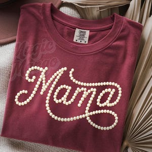 May include: A maroon t-shirt with the word "Mama" spelled out in a cursive font, outlined with small, off-white dots. The words "Magic Mama" are faintly visible above the main text. The shirt is folded and lies on a neutral surface.