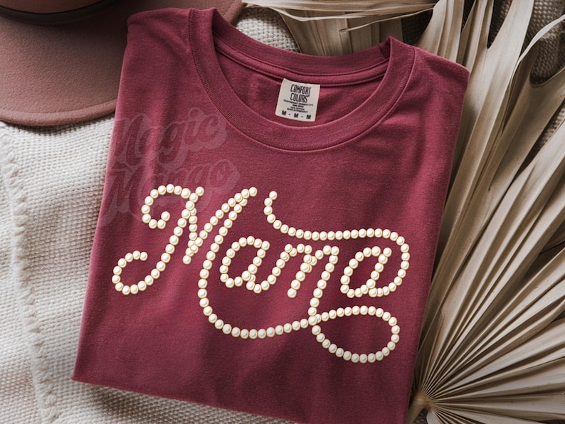 May include: A maroon t-shirt with the word "Mama" spelled out in a cursive font, outlined with small, off-white dots. The words "Magic Mama" are faintly visible above the main text. The shirt is folded and lies on a neutral surface.