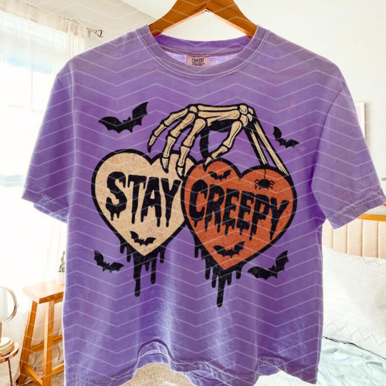 May include: A lilac t-shirt with a Halloween design. The shirt displays two heart-shaped graphics with the words "STAY CREEPY" and a skeleton hand holding them. Bats and dripping details complete the design.