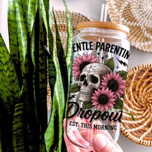 K&ouml;nnte beinhalten: Klare Glaswasserflasche mit Bambusdeckel und Strohhalm. Die Flasche hat ein Totenkopf- und Sonnenblumenmotiv mit dem Text "Gentle Parenting Dropout Est: This Morning".
