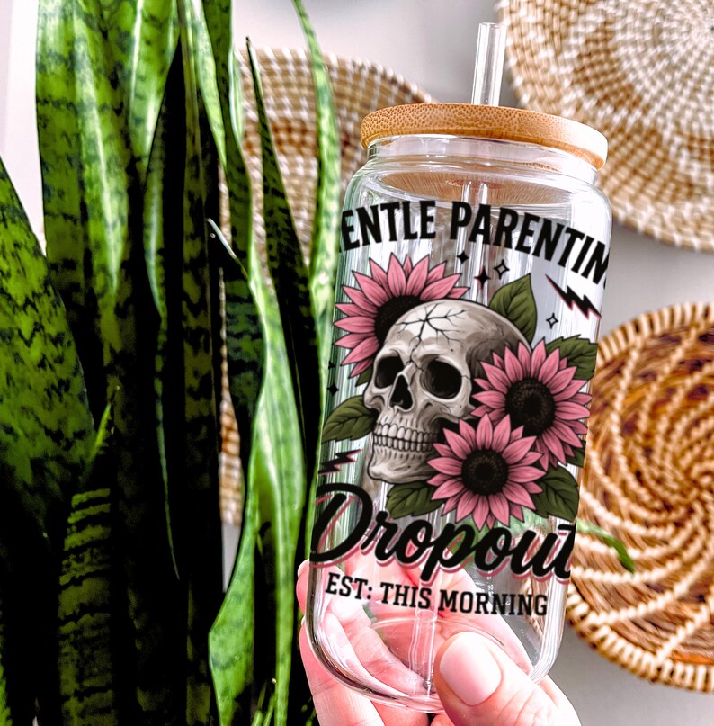 K&ouml;nnte beinhalten: Klare Glaswasserflasche mit Bambusdeckel und Strohhalm. Die Flasche hat ein Totenkopf- und Sonnenblumenmotiv mit dem Text "Gentle Parenting Dropout Est: This Morning".