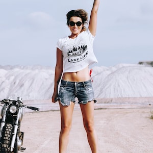 May include: A woman wearing sunglasses, a white crop top with "LAKE LIFE" text, and denim shorts jumps in the air. A vintage motorcycle is parked nearby. The background features a light blue sky and white mounds.