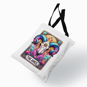 May include: White tote bag with a colourful illustration of a ram skull, featuring blue and purple horns, and floral elements. The text "THE ARIES" is displayed at the bottom. The bag has black handles.