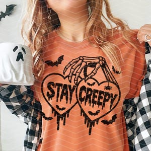 May include: An orange t-shirt with a black graphic. The design features two heart shapes with the words "STAY CREEPY". A skeleton hand holds the hearts, with bats and dripping accents. A white ghost-shaped mug is held in the image.