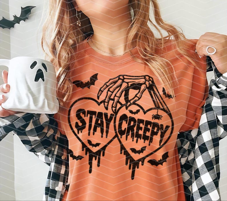May include: An orange t-shirt with a black graphic. The design features two heart shapes with the words "STAY CREEPY". A skeleton hand holds the hearts, with bats and dripping accents. A white ghost-shaped mug is held in the image.