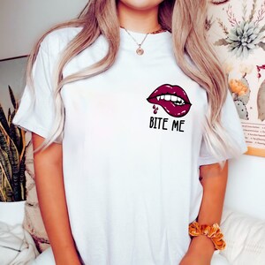 May include: White t-shirt with a red, open-mouthed, vampire-like lip graphic with fangs and dripping blood. The text "Bite Me" is printed below the graphic.