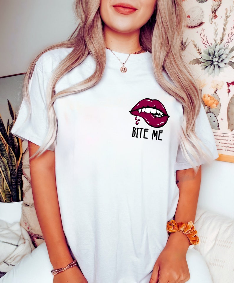 May include: White t-shirt with a red, open-mouthed, vampire-like lip graphic with fangs and dripping blood. The text "Bite Me" is printed below the graphic.
