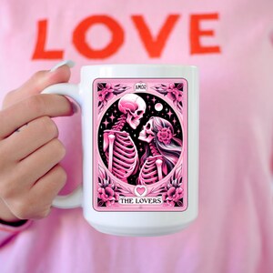 K&ouml;nnte beinhalten: Wei&szlig;e Keramik-Tasse mit einem rosa Tarotkarten-Design mit Skelettfiguren, beschriftet mit "The Lovers" und "AMOR". Die Tasse hat einen gro&szlig;en Henkel. Die Person, die die Tasse h&auml;lt, tr&auml;gt ein rosa T-Shirt mit dem Wort "LOVE" in Rot.