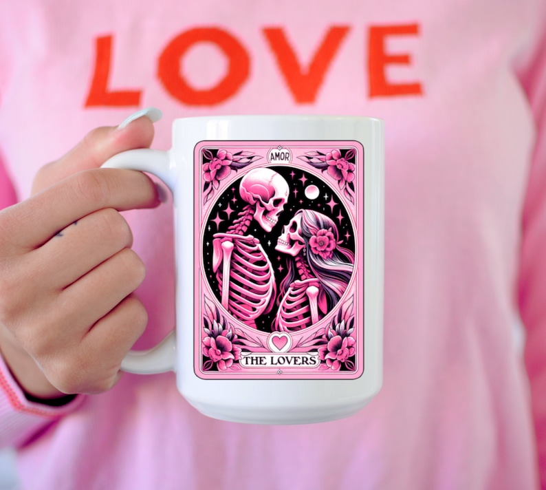 K&ouml;nnte beinhalten: Wei&szlig;e Keramik-Tasse mit einem rosa Tarotkarten-Design mit Skelettfiguren, beschriftet mit "The Lovers" und "AMOR". Die Tasse hat einen gro&szlig;en Henkel. Die Person, die die Tasse h&auml;lt, tr&auml;gt ein rosa T-Shirt mit dem Wort "LOVE" in Rot.