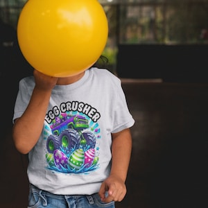 May include: A child wearing a white t-shirt with a graphic of a monster truck driving over Easter eggs. The text "Egg Crusher" is written in a colourful arc above the truck. The child is holding a yellow balloon in front of their face.