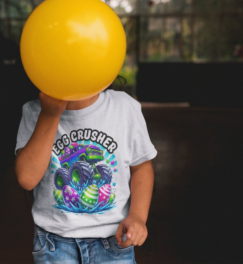 May include: A child wearing a white t-shirt with a graphic of a monster truck driving over Easter eggs. The text "Egg Crusher" is written in a colourful arc above the truck. The child is holding a yellow balloon in front of their face.
